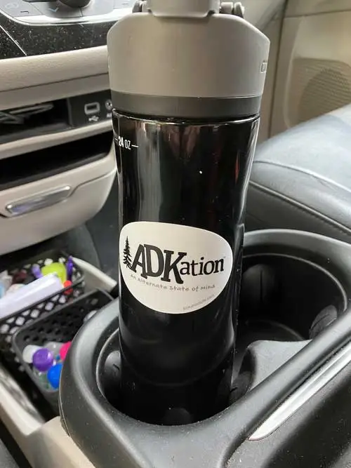Black travel mug in a car cup holder with white ADKation™ sticker. Example of a unique Adirondack vacation souvenir.