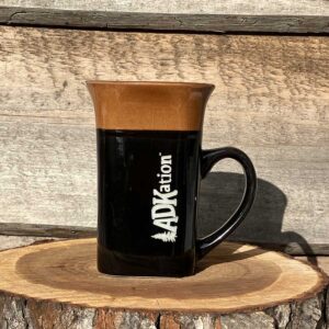 A tall brown and black coffee ADK vacation souvenir mug for right hand drinkers with the text “ADKation” vertically engraved near the handle, feeding memories of fun Adirondack vacation adventures and experiences.