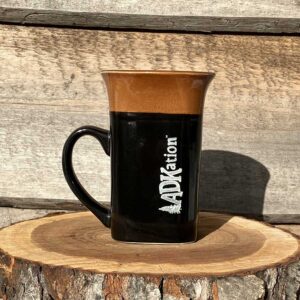 A tall brown and black coffee ADK vacation souvenir mug for left handers with the text “ADKation” vertically engraved near the handle, keeping left handers energized with memories of Adirondack vacation smiles and memories.