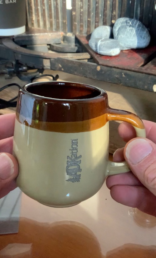 A two tone, old style ADK souvenir coffee mug with the text “ADKation” engraved vertically to fuel your Adirondack vacation senses, at home, camp, workspace, or wherever you find yourself.