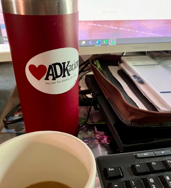 ADK vacation merchandise for your everyday, anywhere connection to Adirondack vacation moments. At home, cottage, cabin, or as in this image, your work space. Features a water bottle on a desk with a sticker that includes an image of a pine tree followed by the text ‘ADKation’ and a tag line of ‘An alternate state of mind.’