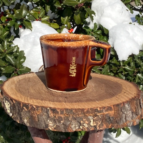 Adirondack Christmas Stocking Stuffer | Vintage Hull Pottery brown drip coffee mug with ‘ADKation’ engraved vertically near the handle, with snow and a holly tree in the background. | A meaningful Christmas gift idea.